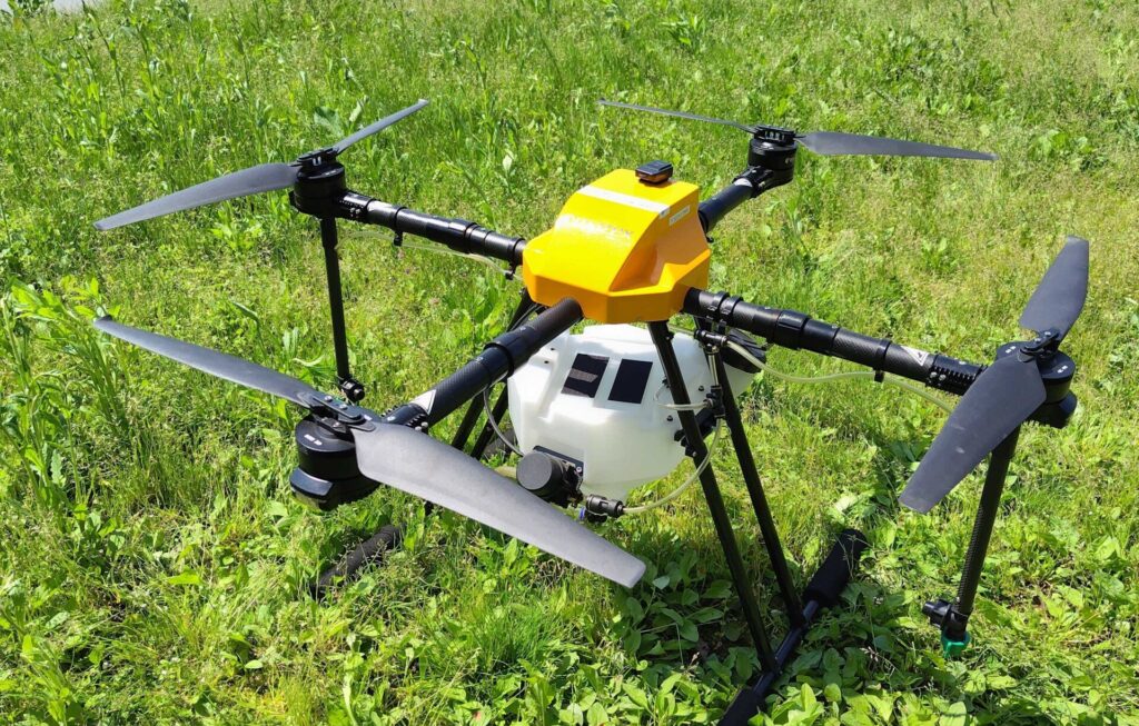 農地の草地に設置された大型農業用ドローン。黄色いトップカバーと白い薬剤タンクを備えており、液体散布用ノズルが機体下部に取り付けられている。背景には水田、ビニールハウス、農業施設が広がっている。